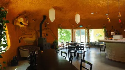 Gastraum im Erdhaus mit einer plastischen Drachenfigur an der Wand, Holzofen und langem Tisch