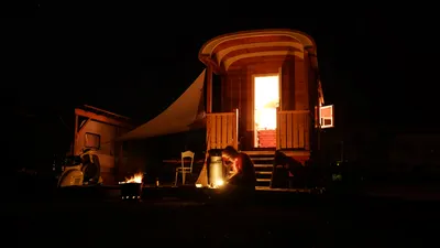Beleuchteter Zirkuswagen bei Nacht, davor jemand am Feuer
