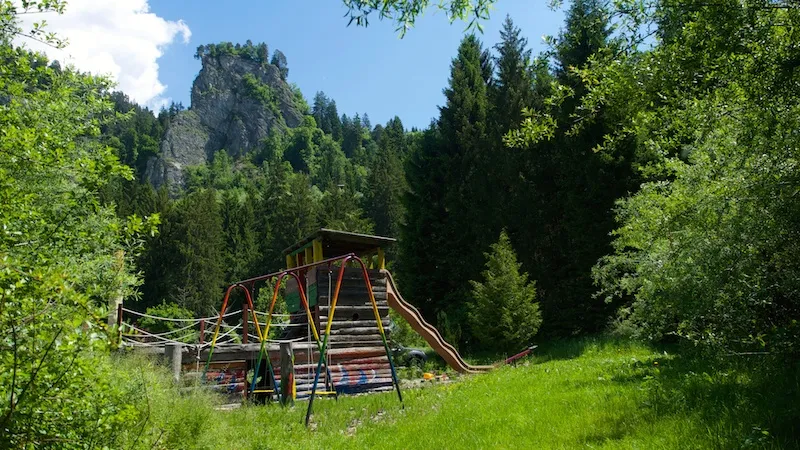 Spielplatz mit Holzturm, Rutsche und Schaukel, dahinter der Felsturm über dem Wald
